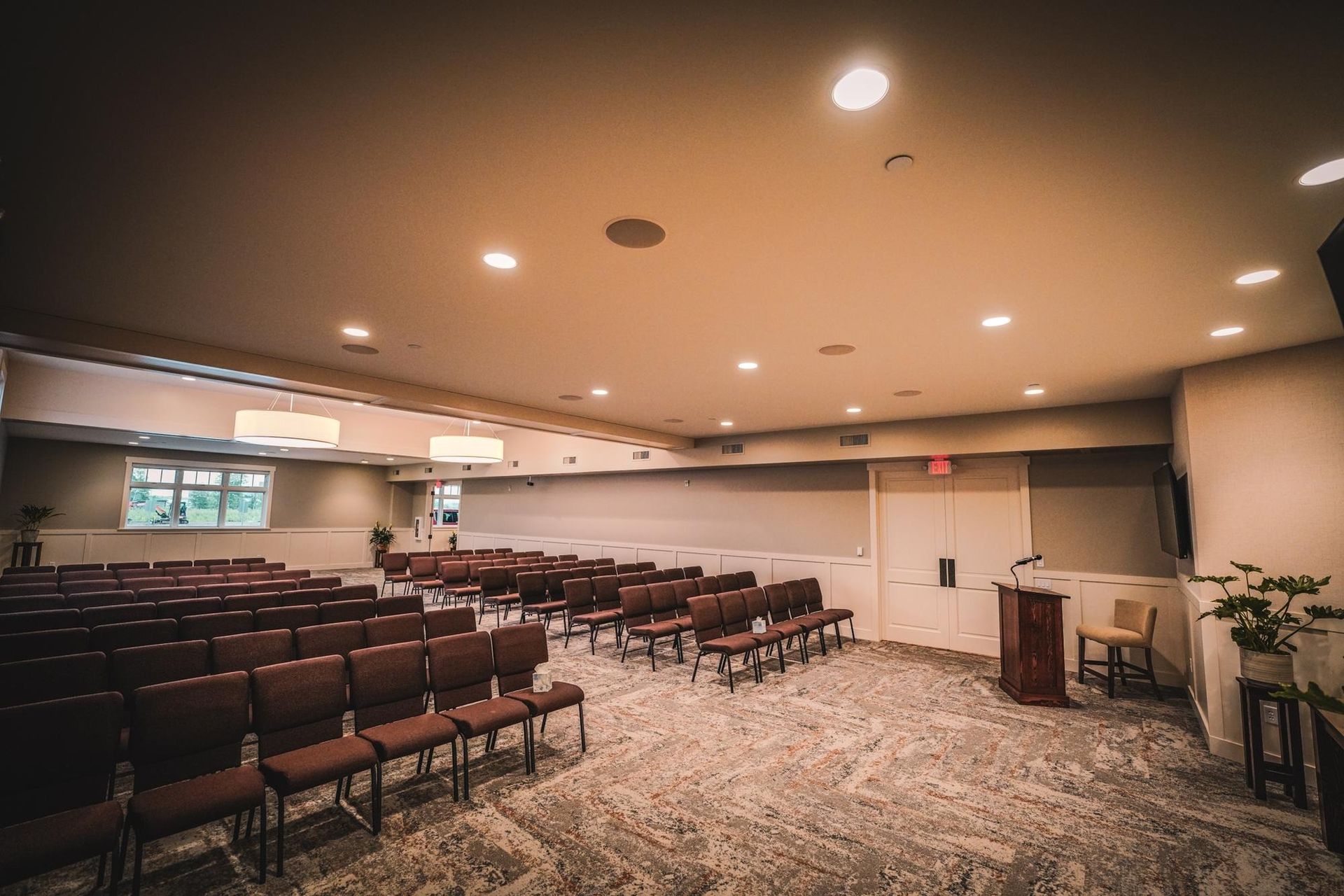 Rows of empty chairs face a wooden podium in a carpeted room with neutral tones, likely a meeting space.