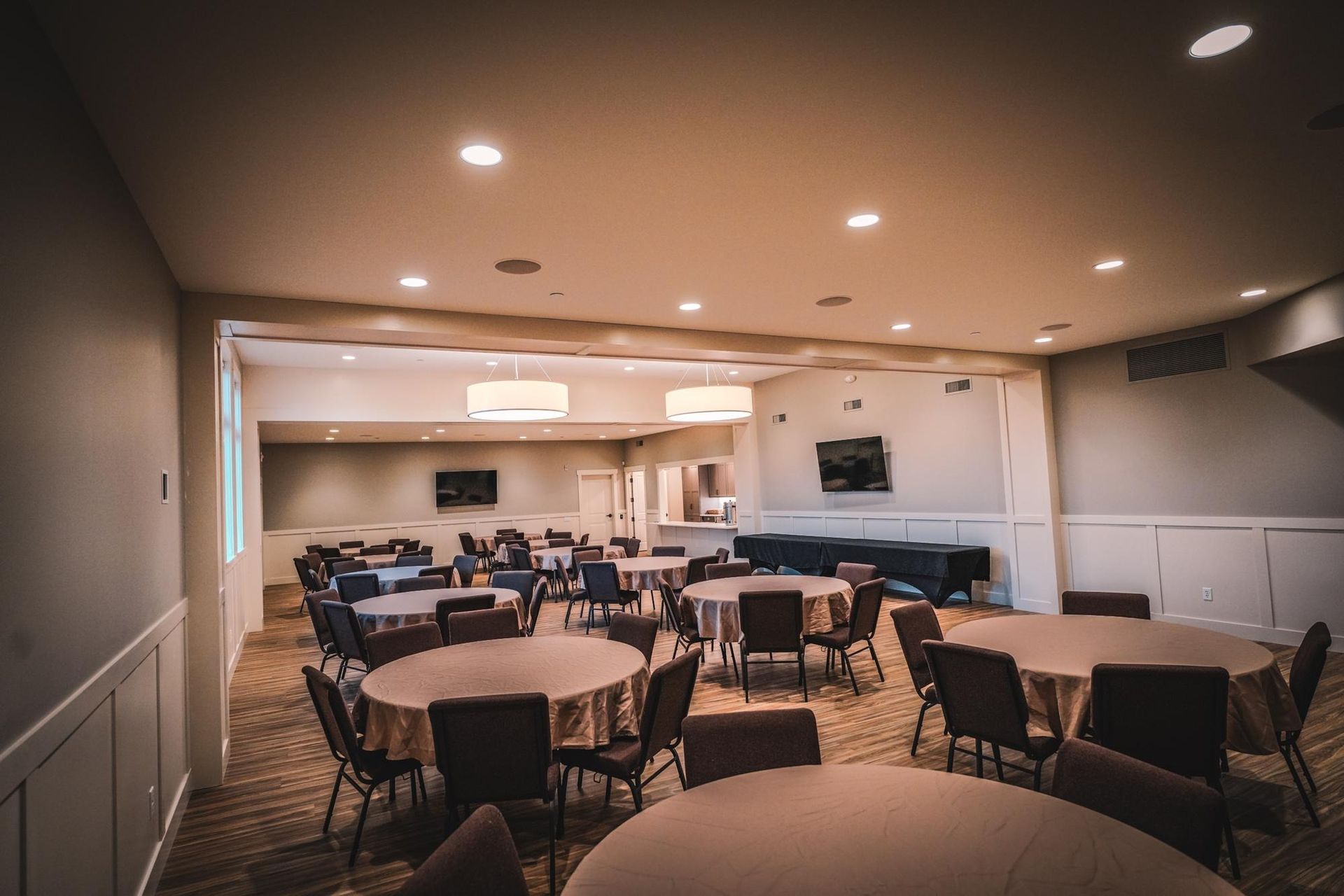 Event room with round tables draped in tan cloths, chairs, and recessed lighting.