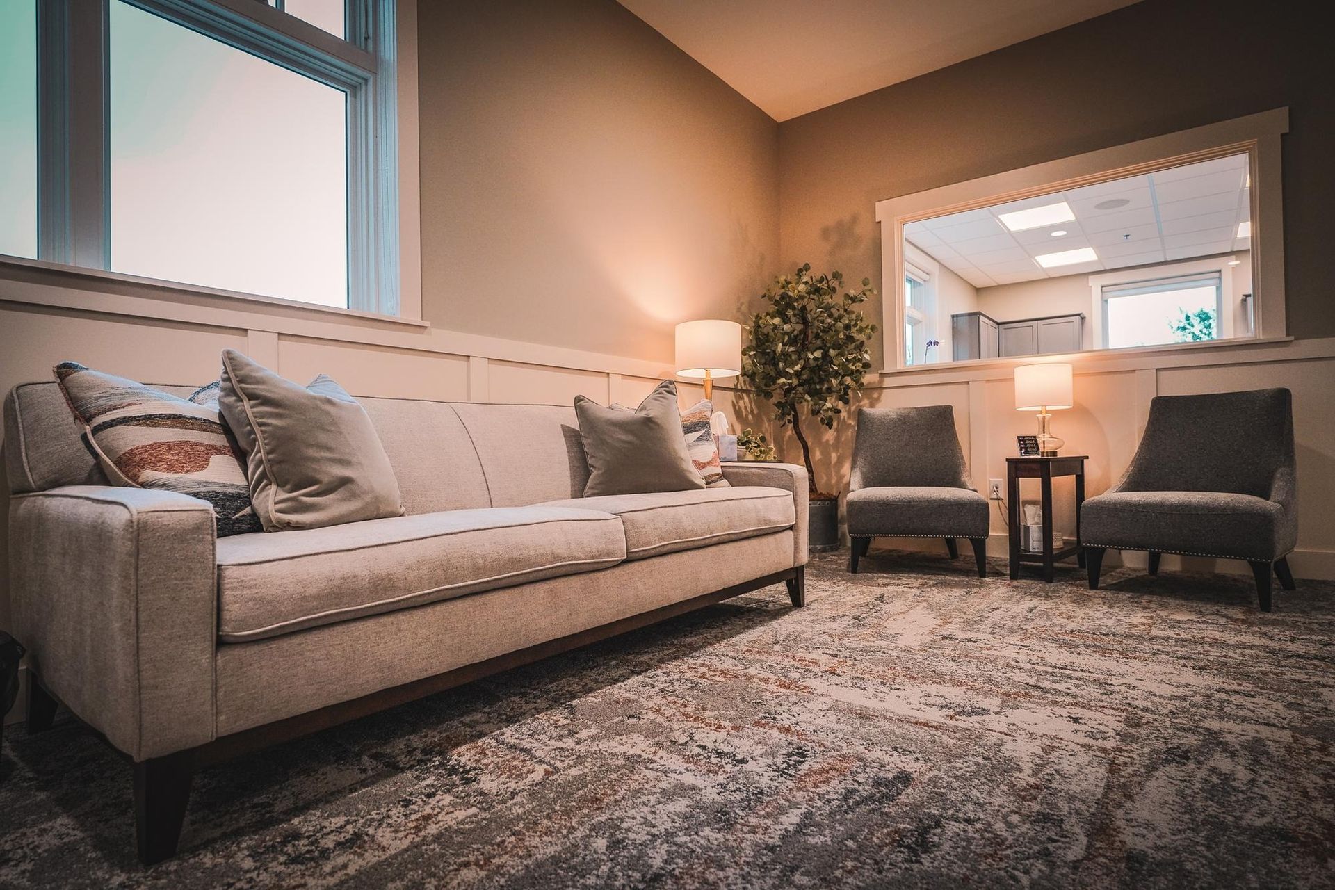 Living room with beige sofa, two chairs, and a window. Neutral tones, rug, and lamps.