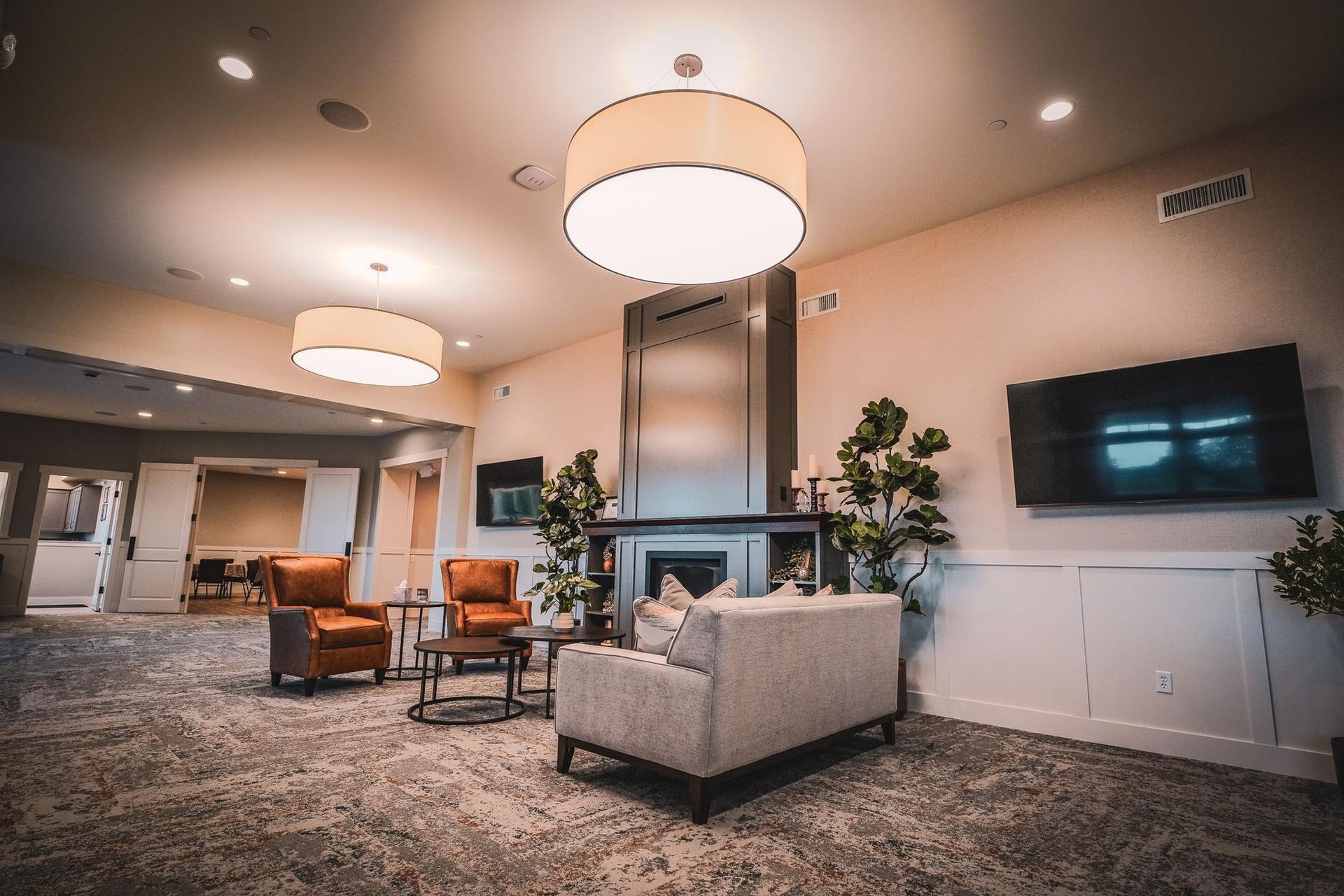 Lounge area with a fireplace, seating, two TVs, and two large pendant lights. Neutral tones, carpeted floors.