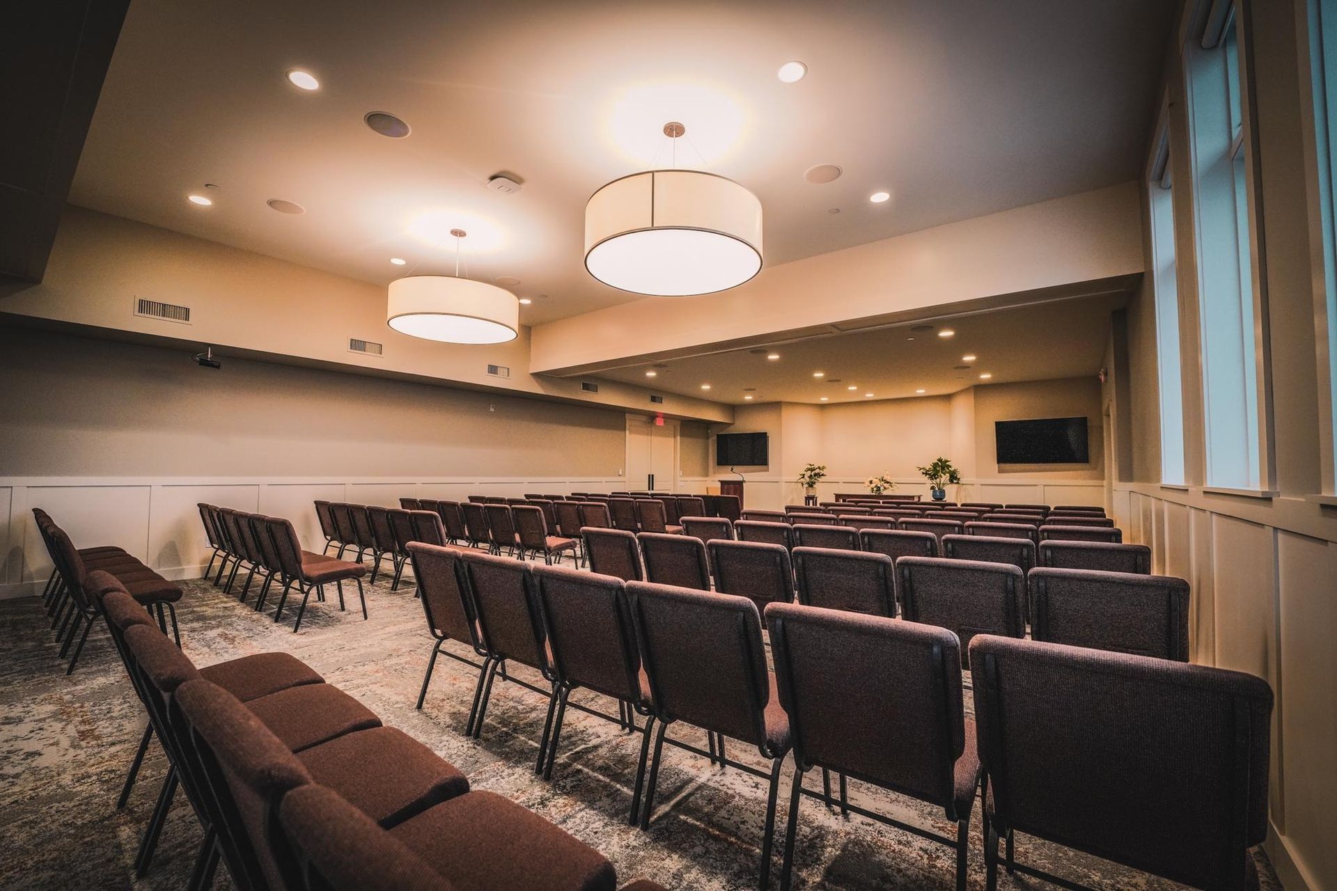 Rows of empty chairs face a stage in a beige-walled room, two large lights hang from the ceiling.