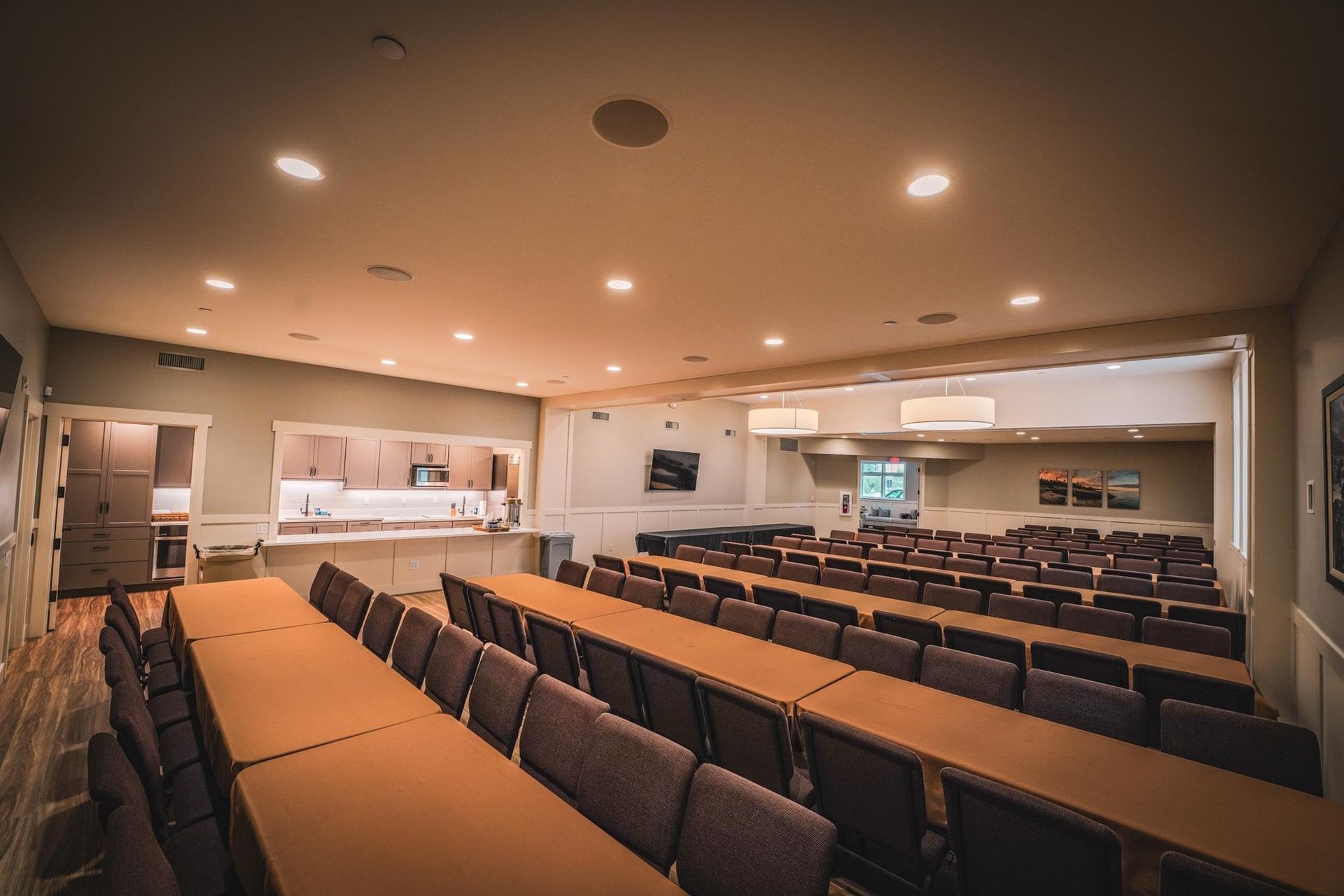 Empty event space with rows of tables and chairs, a small kitchen area, and overhead lighting.