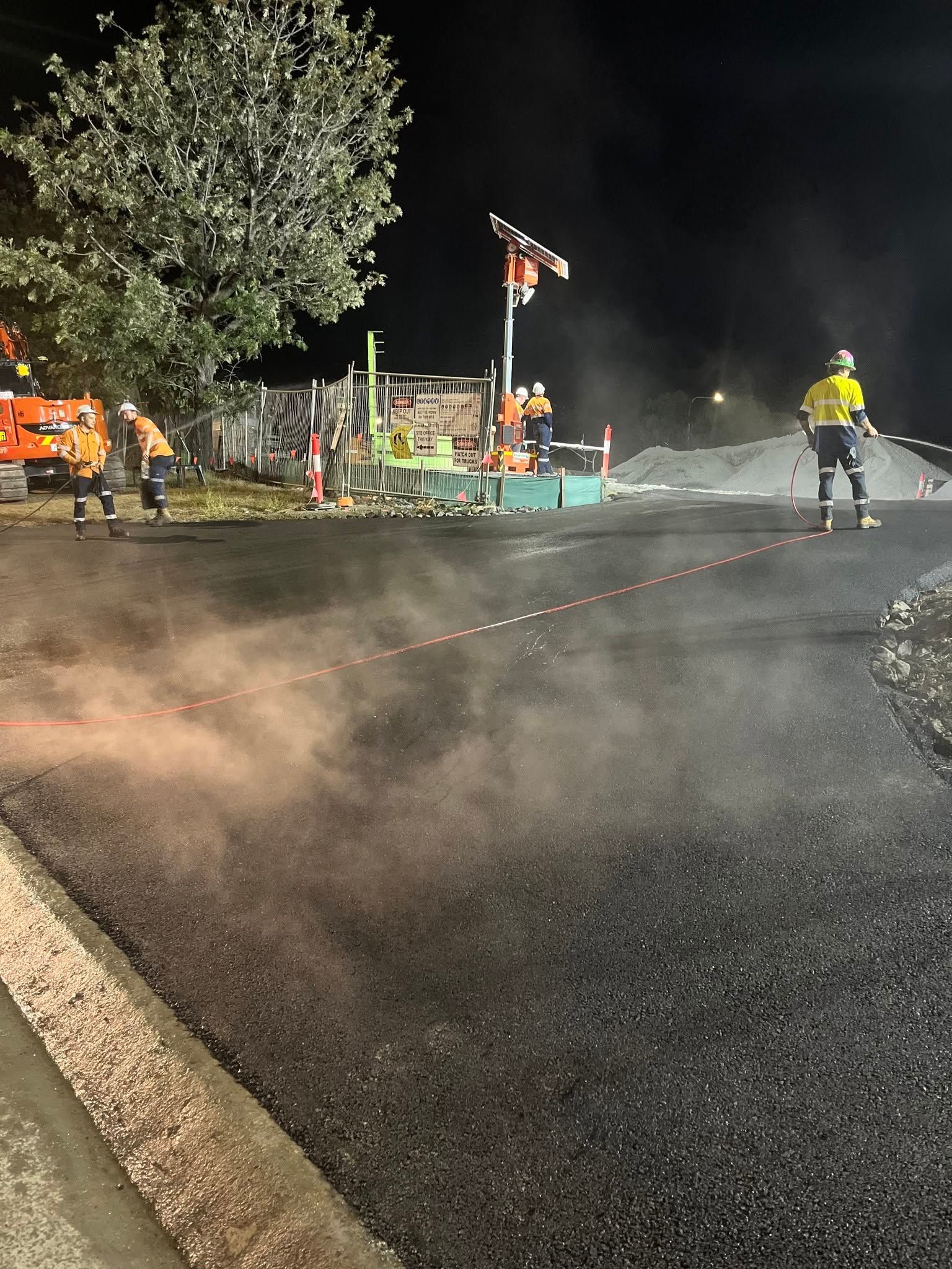 Night roadwork scene with workers, steam rising from wet pavement, and a tree lit by work lights.