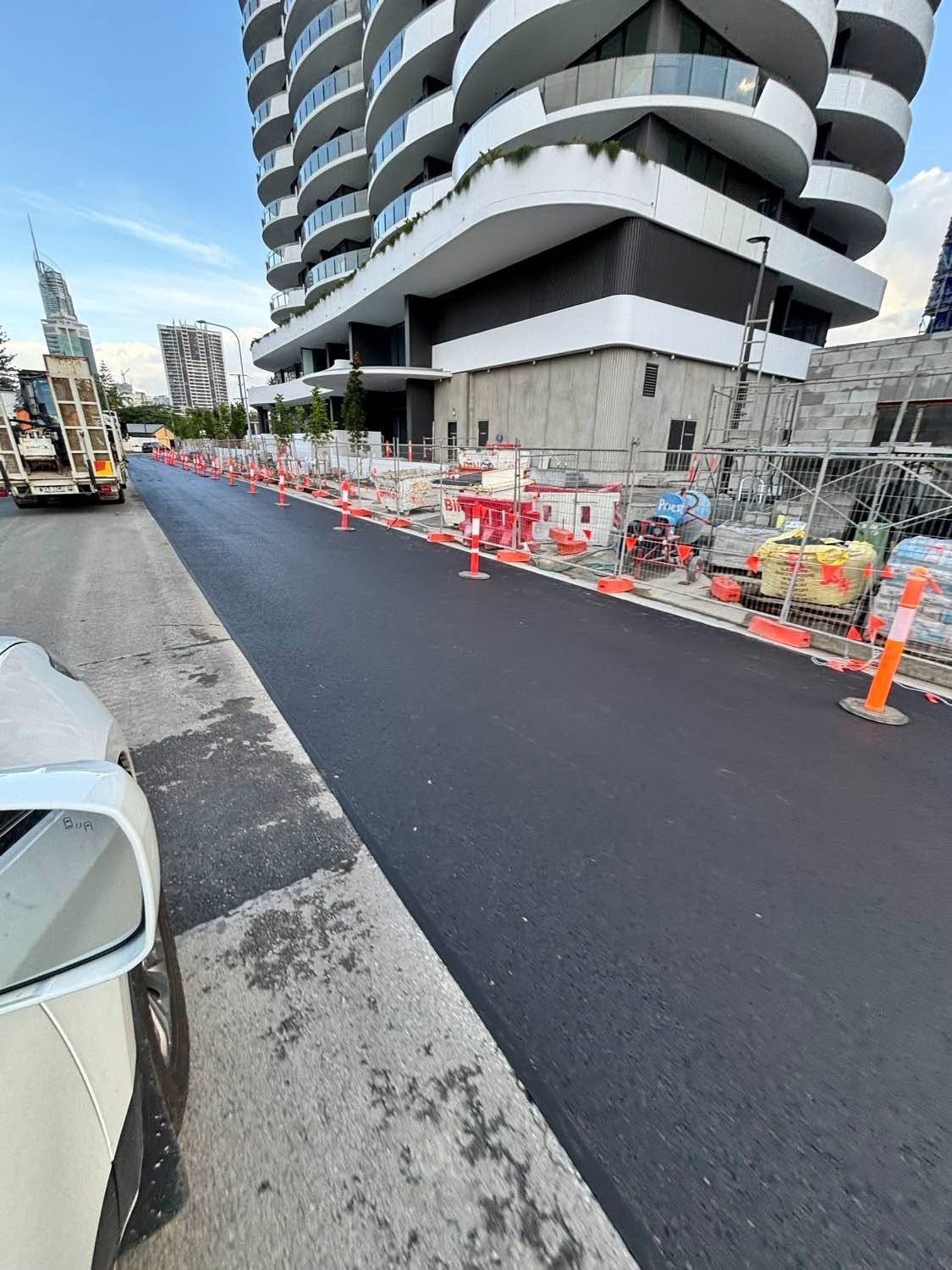 Roadworks beside a curved apartment building, with orange cones and barriers lining a newly paved street.