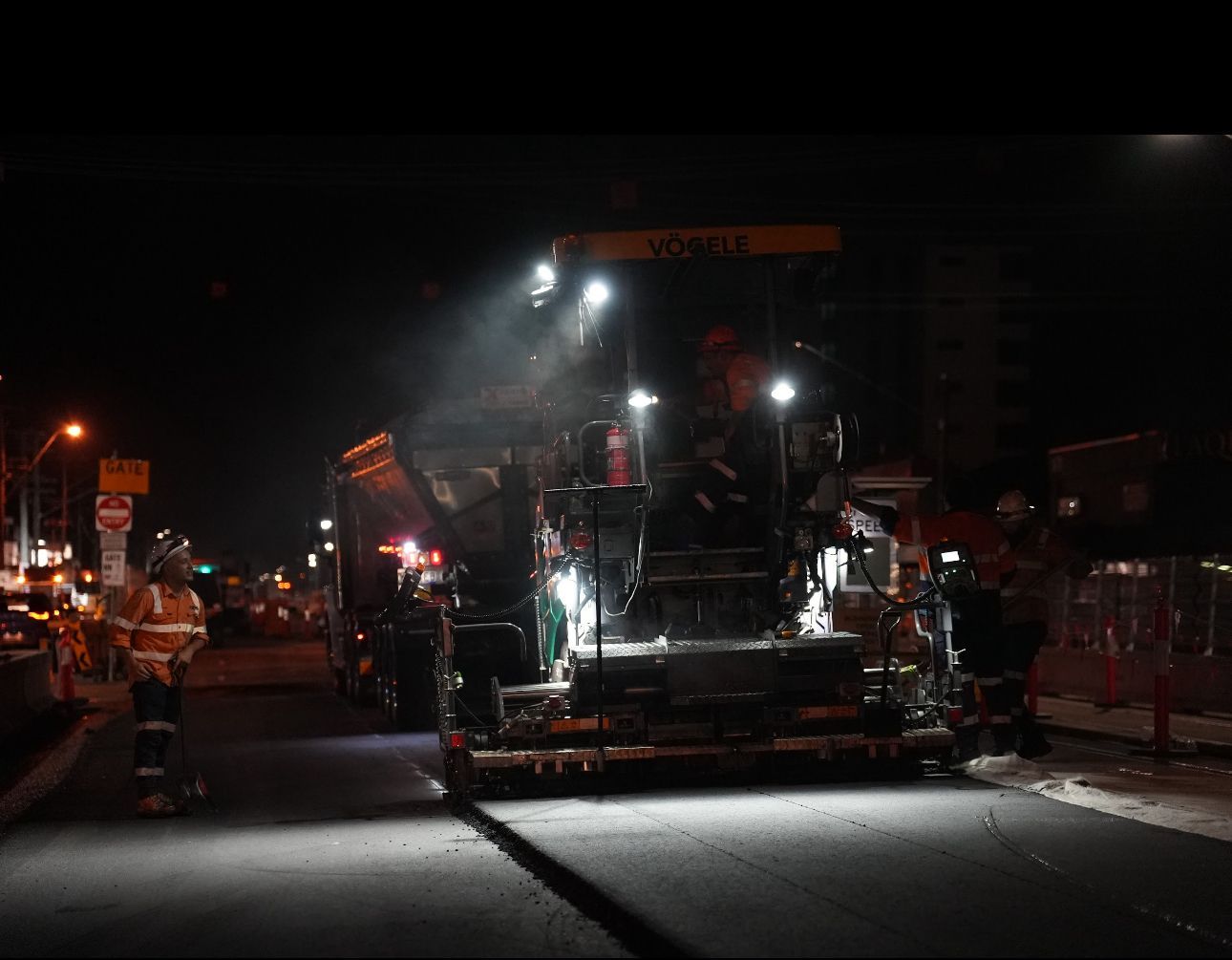 Asphalt paving machine illuminated at night, laying pavement. Workers in safety vests nearby.