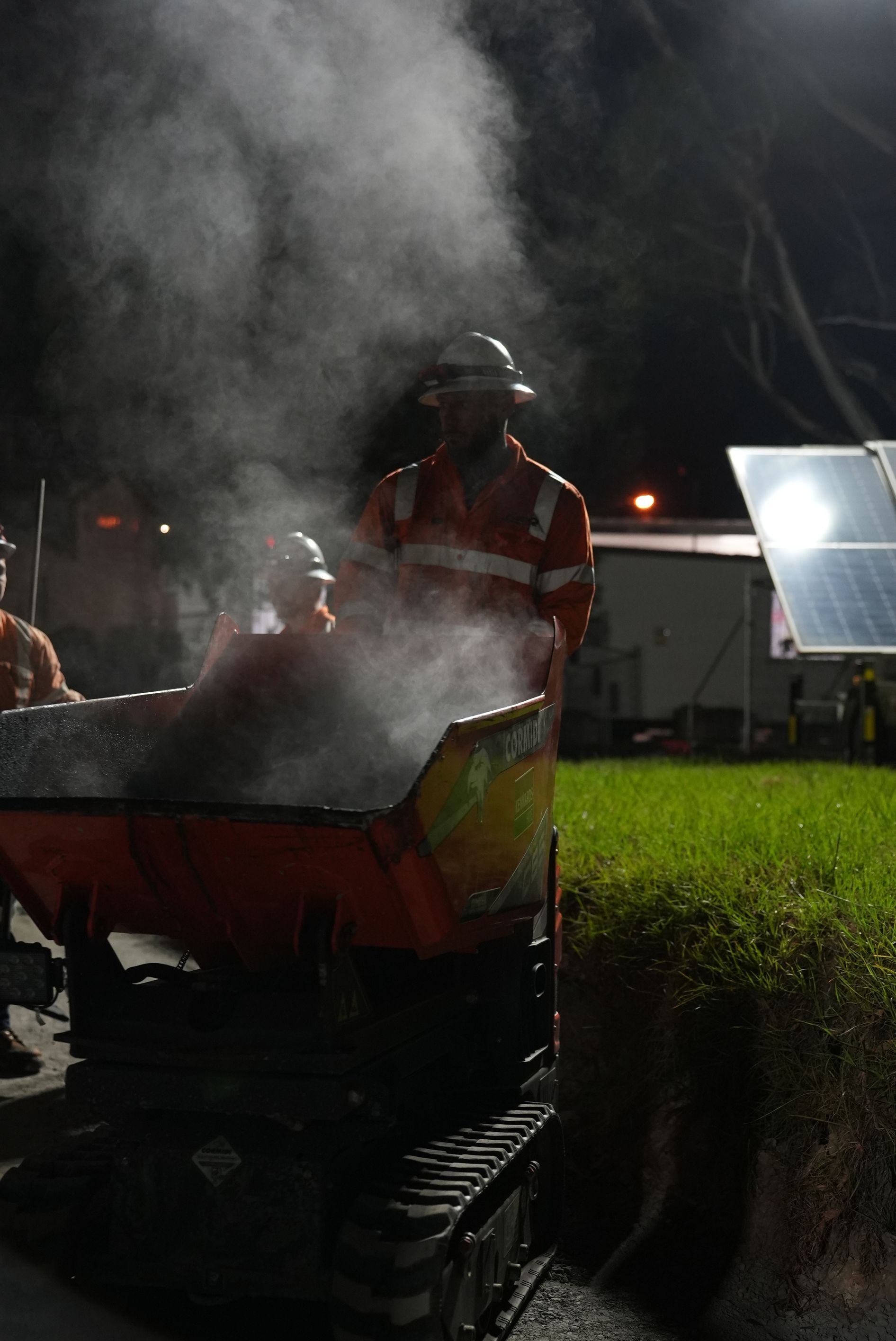 Workers in orange safety vests near steaming asphalt equipment at night.