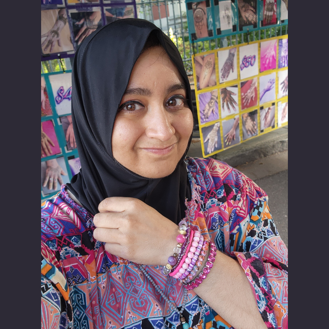 Smiling person in a black hijab and colorful dress, holding a hand near their chin in front of patterned artwork.