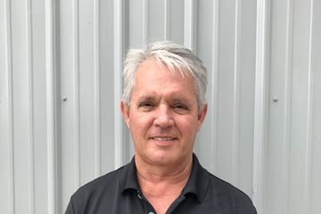 Portrait of a smiling person with gray hair, wearing a black shirt, standing before a corrugated metal wall