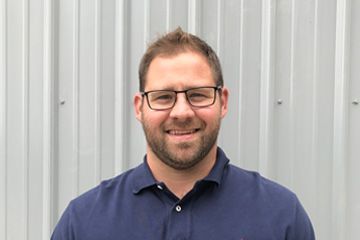 Smiling man with glasses in a blue shirt standing against a light gray metal wall