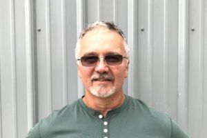 Man wearing sunglasses and a green shirt standing in front of a corrugated metal wall