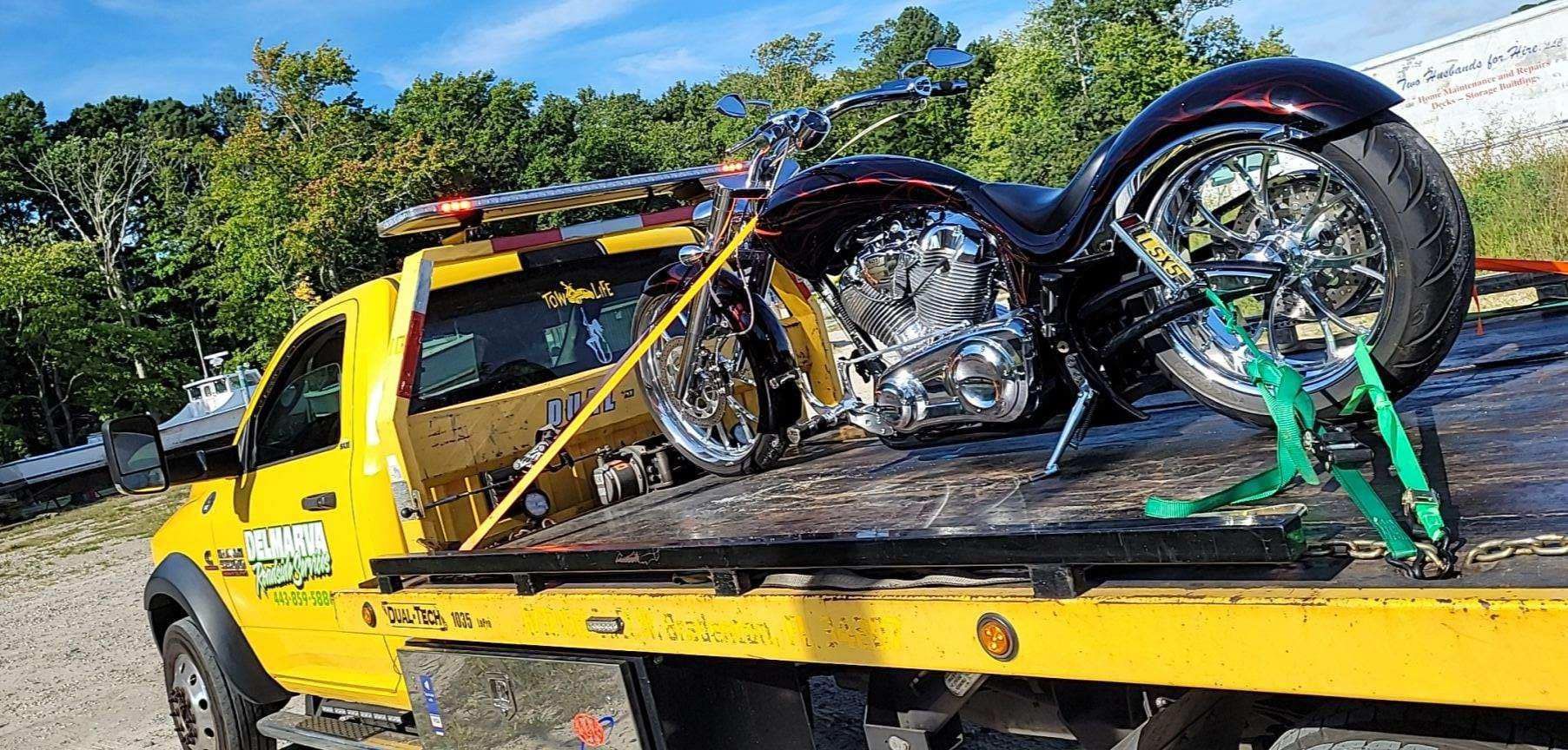 Yellow tow truck carrying a crushed motorcycle on a flatbed roadside, with trees in the background