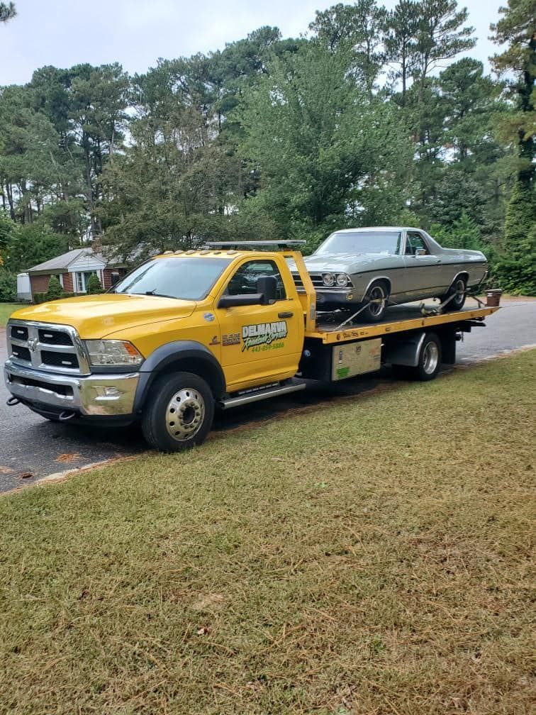 Yellow tow truck carrying a gray classic car on a flatbed beside a grassy roadside