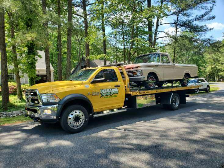 Yellow tow truck carrying a beige pickup on a flatbed in a wooded driveway