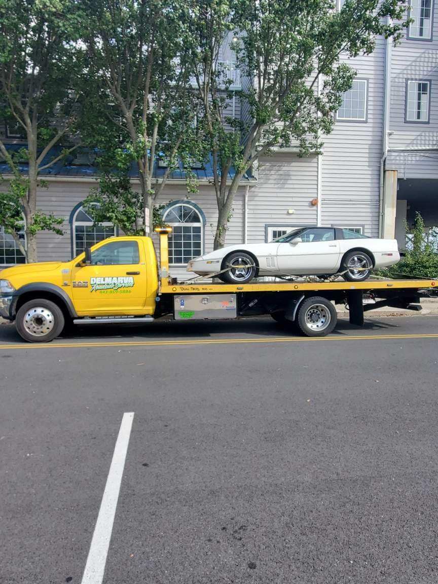 Yellow tow truck carrying a white sports car parked on a city street with apartment buildings behind.