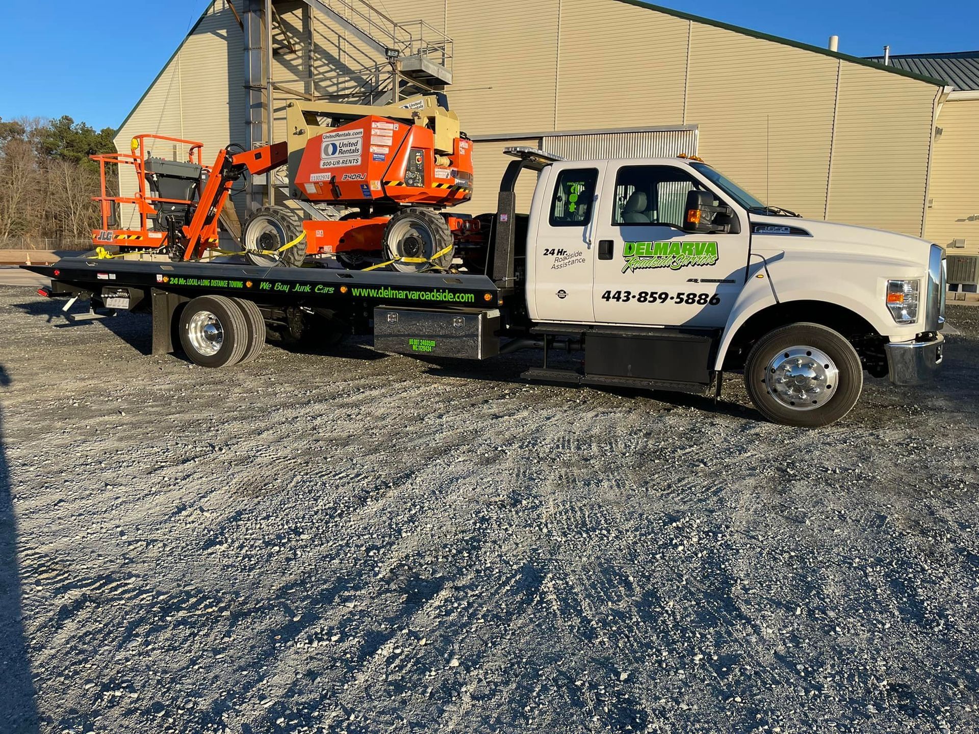 White flatbed tow truck carrying orange mini excavators in a gravel lot beside a beige building.