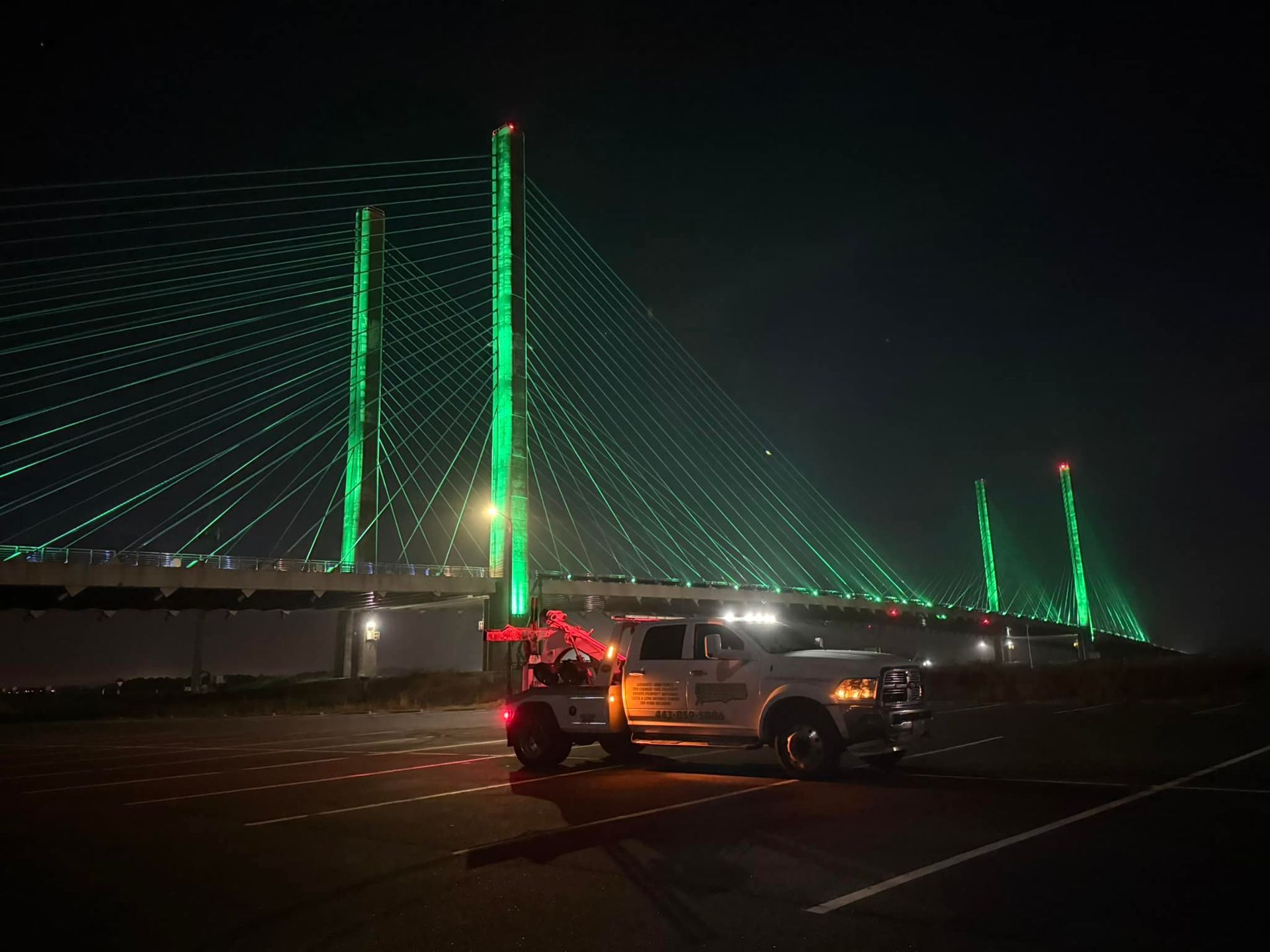Night scene of a lit suspension bridge with green lights and a pickup truck towing a motorcycle in the foreground