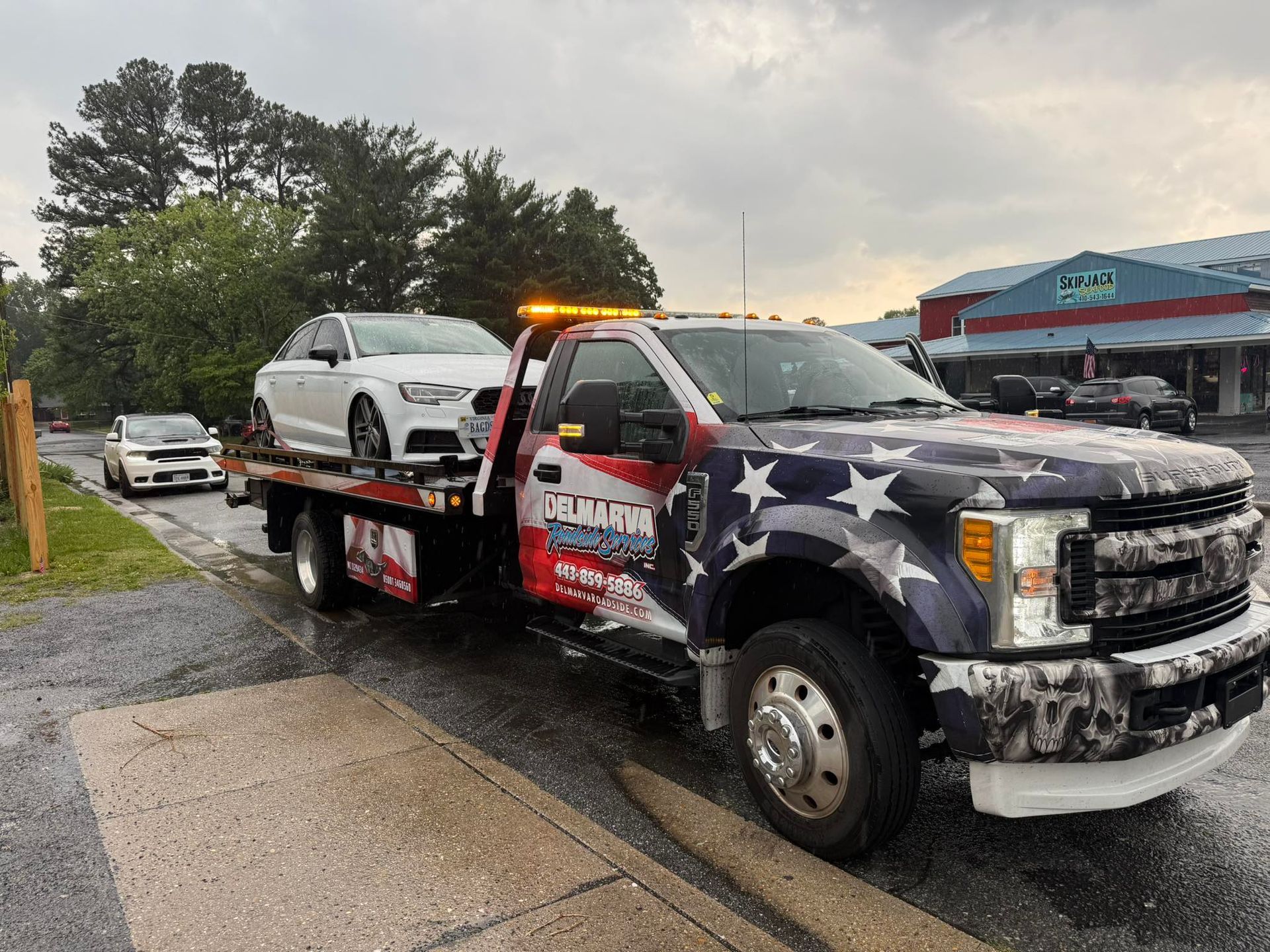 Tow truck carrying a white SUV on a rainy roadside, with an American flag design on the truck.