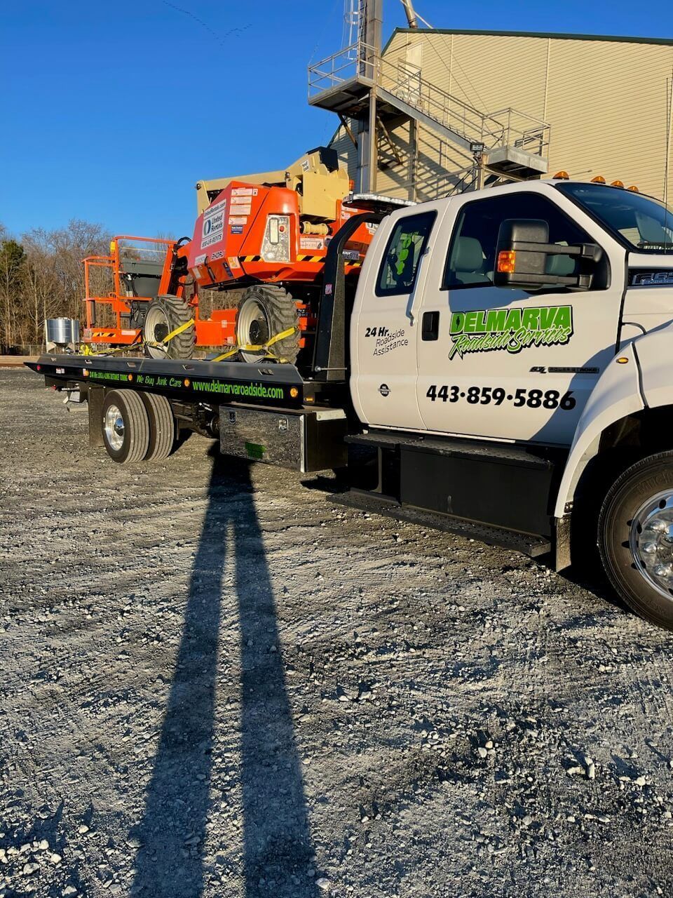 White flatbed tow truck carrying orange machinery on a gravel lot under a clear blue sky