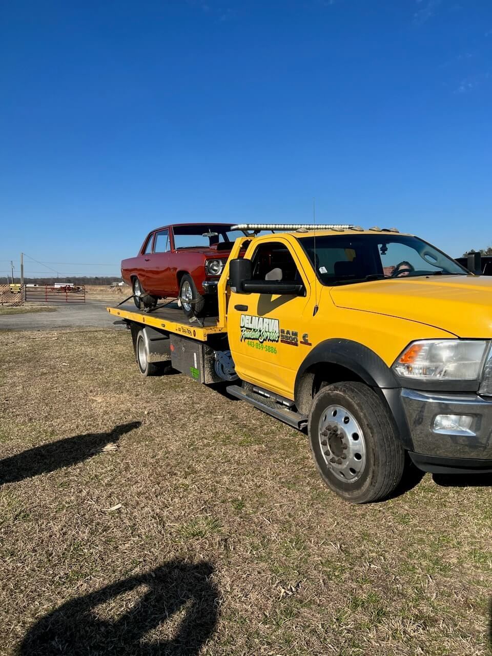 Yellow tow truck carrying a red pickup on a flatbed in a sunny gravel lot