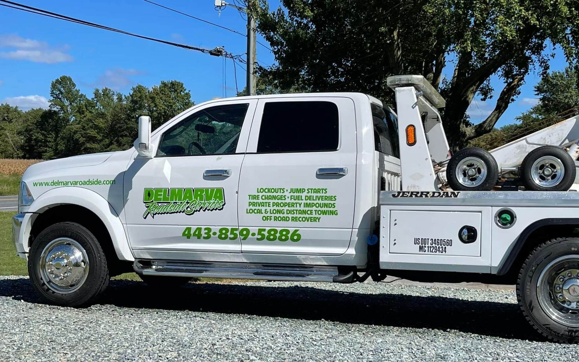 White tow truck with green lettering and a car on the back, parked outdoors on a sunny day.