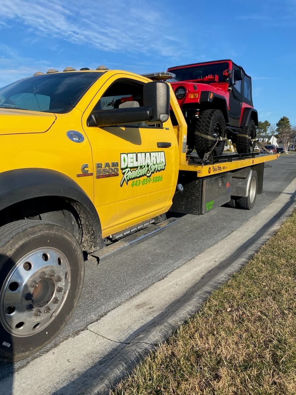 Yellow tow truck hauling a red Jeep on a flatbed beside a road