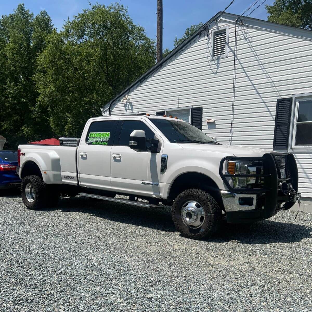 White pickup truck with utility rack parked on gravel beside a white house on a sunny day