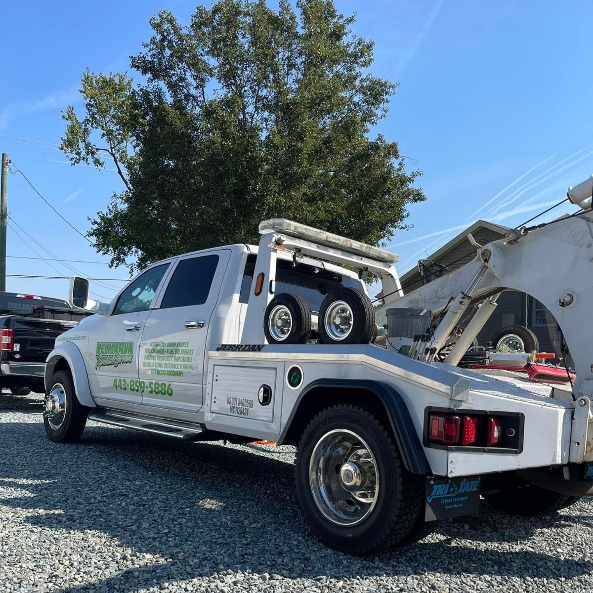 White tow truck parked on gravel, side view with equipment bed, under a tree on a sunny day.