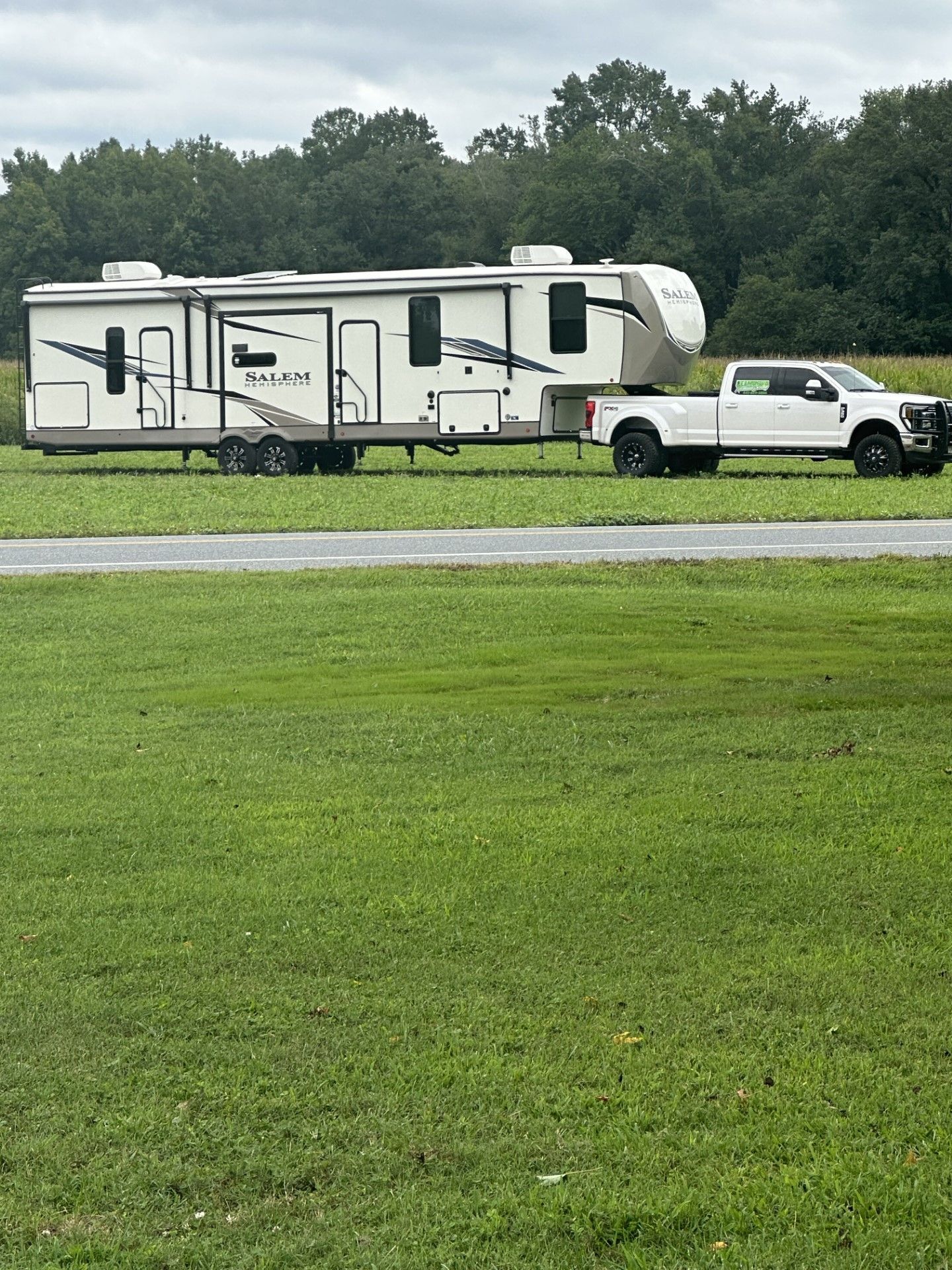 White pickup towing a cream RV trailer beside a wet road in a grassy field, under cloudy skies