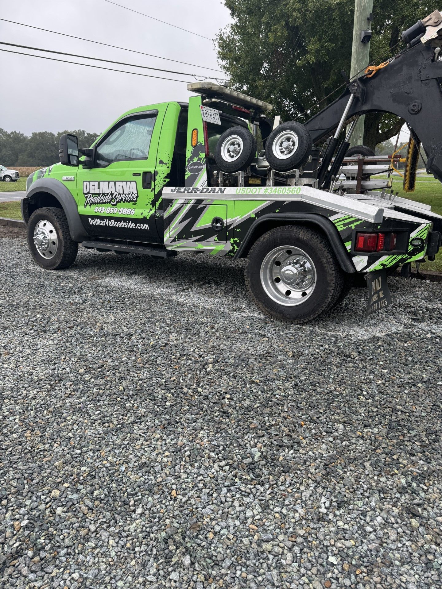 Green and black tow truck parked on gravel, carrying a small vehicle loader with a parked car in the back.