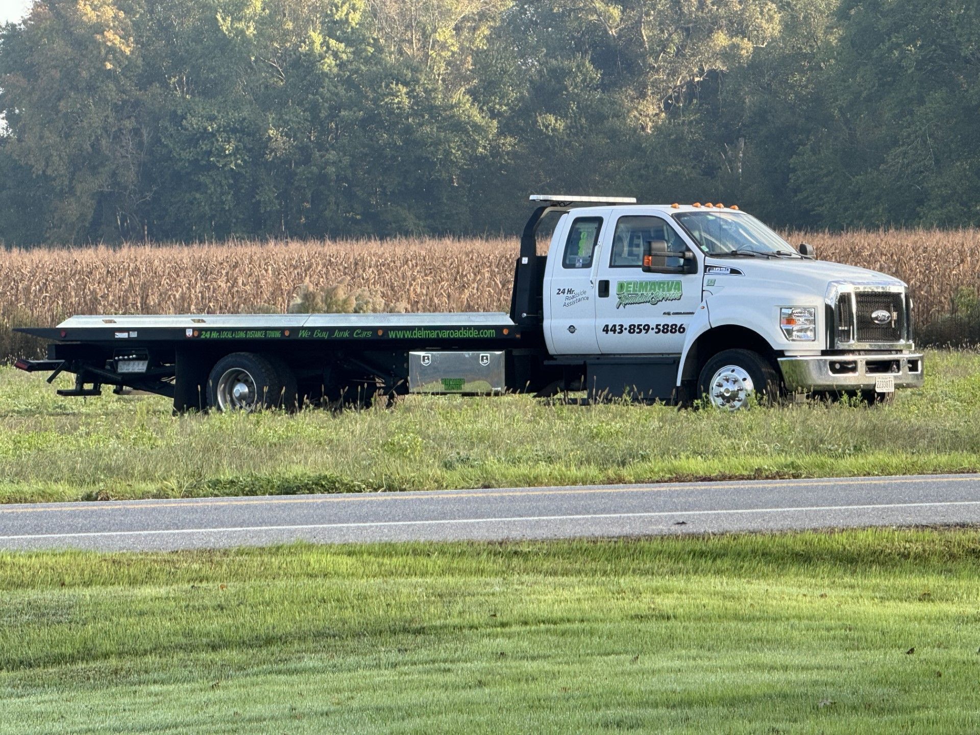 White flatbed tow truck parked by a cornfield on a rural road.
