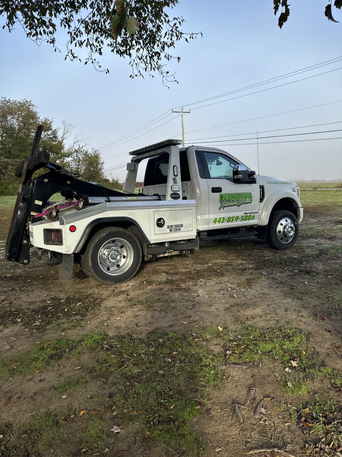 White flatbed tow truck parked on a dirt slope under trees with power lines overhead