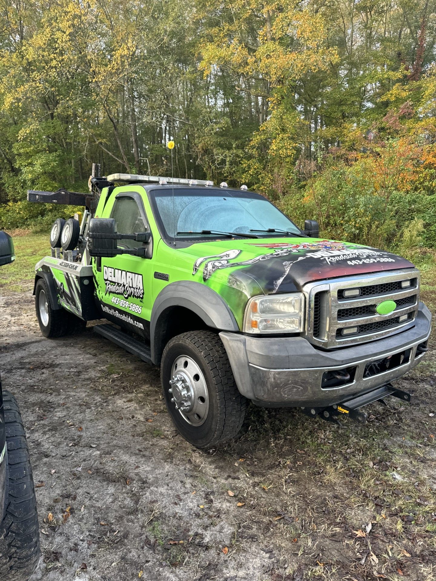 Bright green off-road tow truck parked on muddy ground in a wooded area, autumn trees behind it