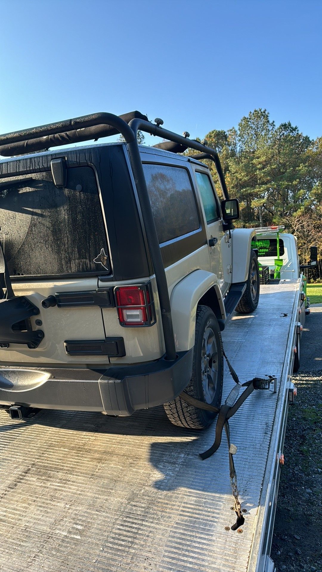 Damaged silver SUV on a tow truck beside a wooded road, rear window shattered and debris visible.