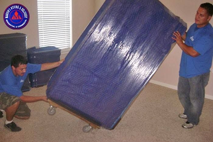 Two men in blue shirts moving a large blue mattress through a room