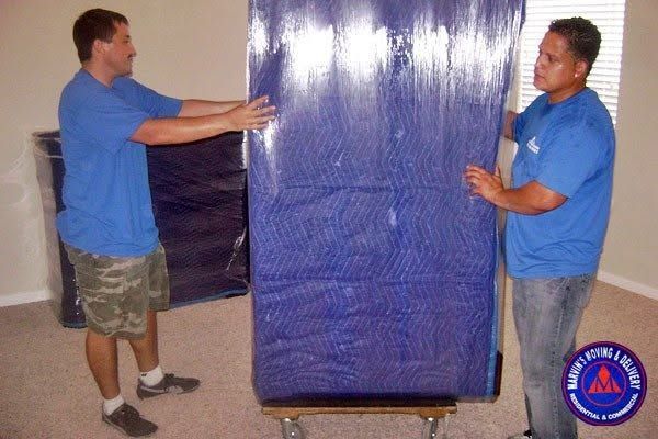 Two men in blue shirts lifting a large wrapped object on a rolling cart in a room