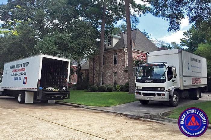 Two white moving trucks parked by a brick house on a tree-lined street