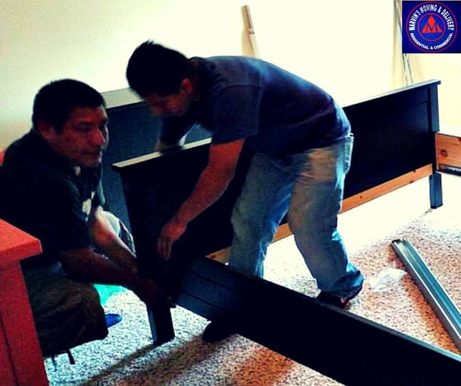 Two men assembling a black metal bed frame in a room with carpet and a wooden dresser