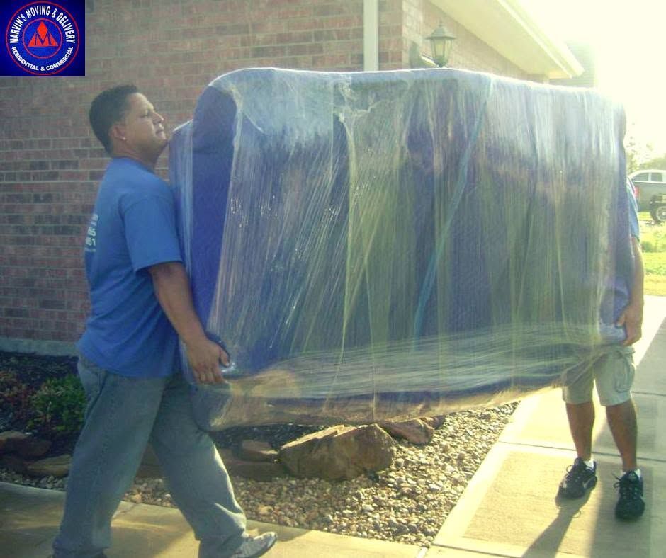 Two men carrying a large wrapped appliance beside a brick building in bright sunlight