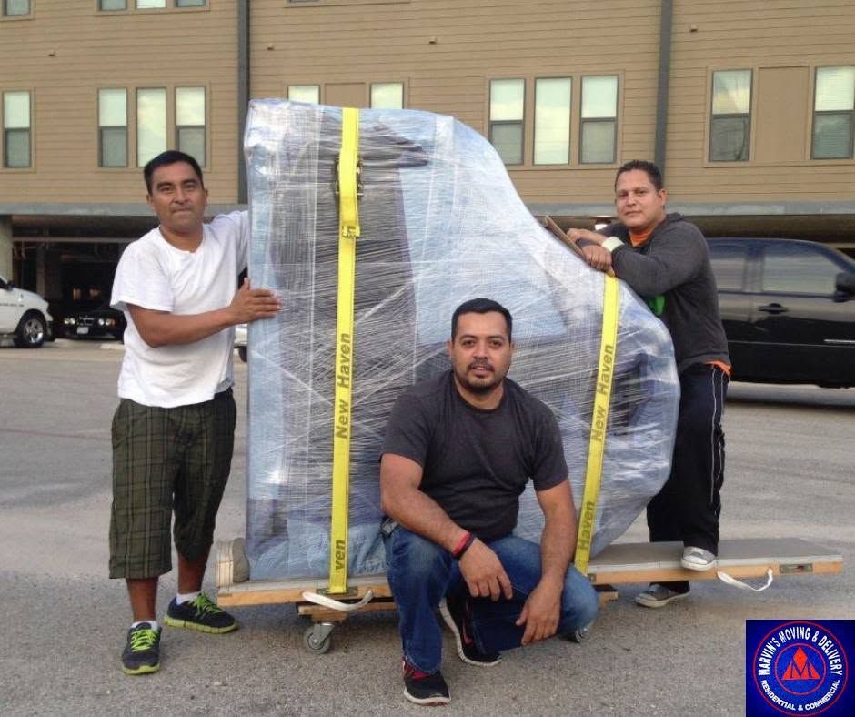 Three men outside a building pose with a large wrapped object strapped to a dolly with yellow straps.