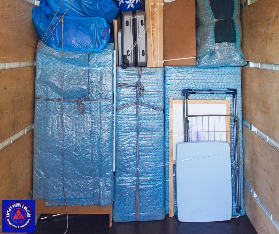 Packed blue-wrapped furniture and boxes stacked in a storage unit, with a folded white chair in front