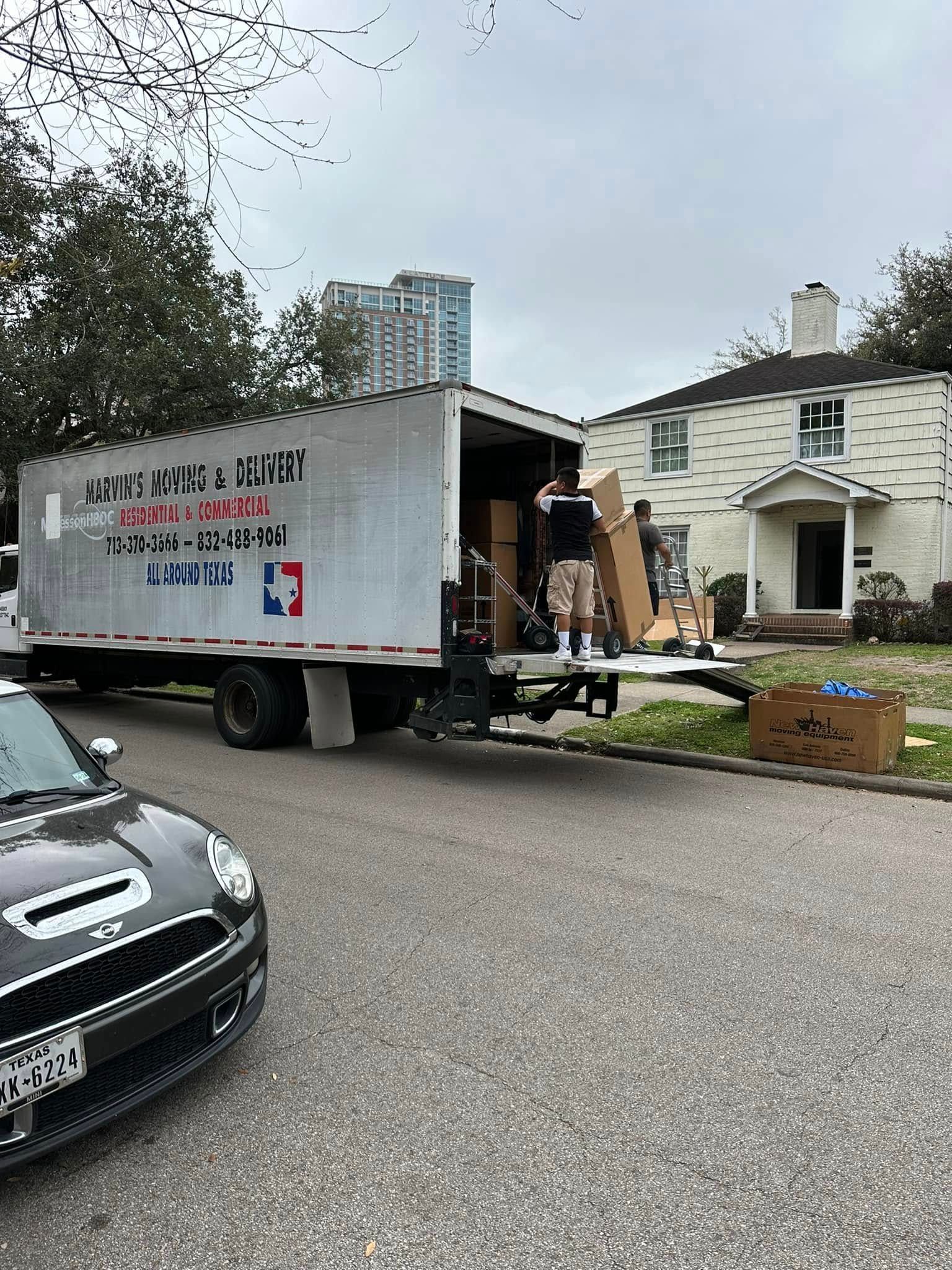 Moving truck parked beside a house, with workers unloading furniture and boxes into the driveway.