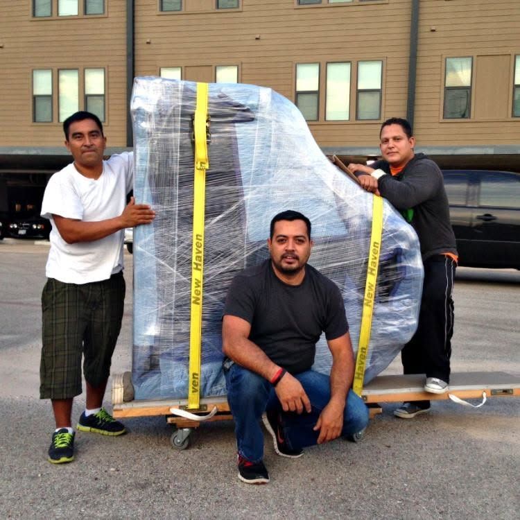 Three men pose beside a large wrapped object on a pallet in a parking lot