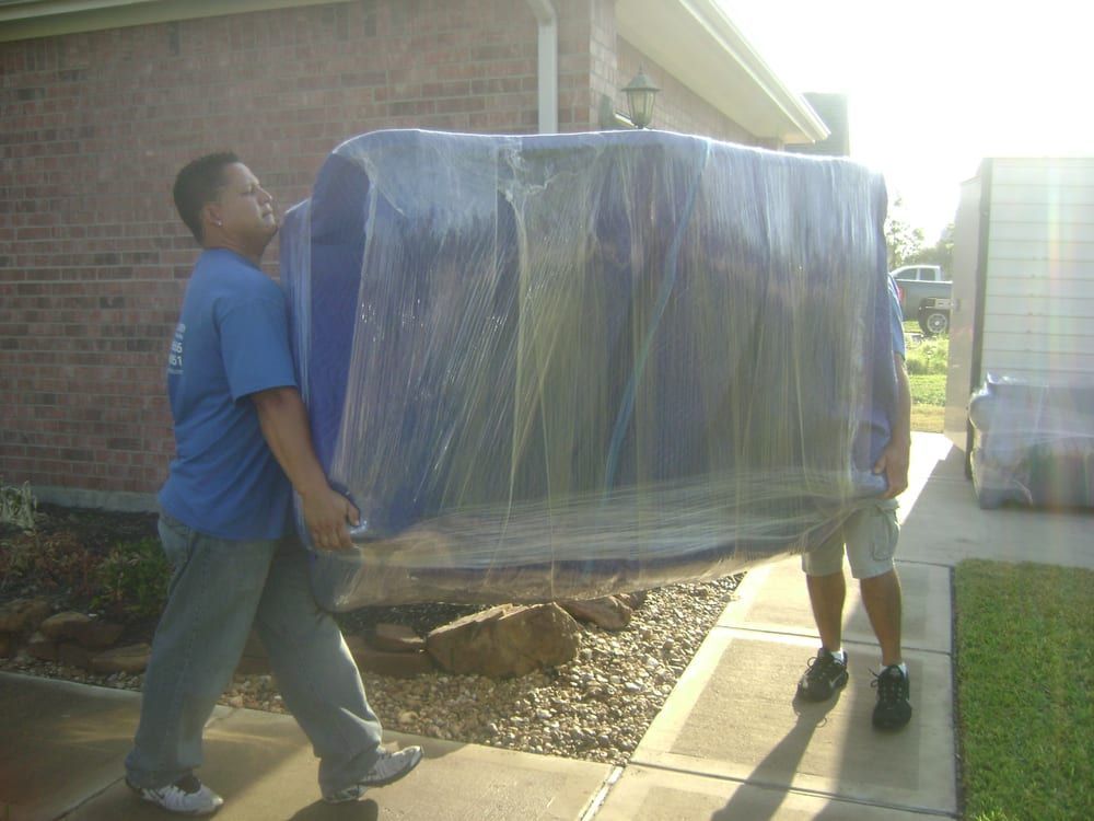 Two people carrying a large wrapped appliance on a dolly outside a brick building.
