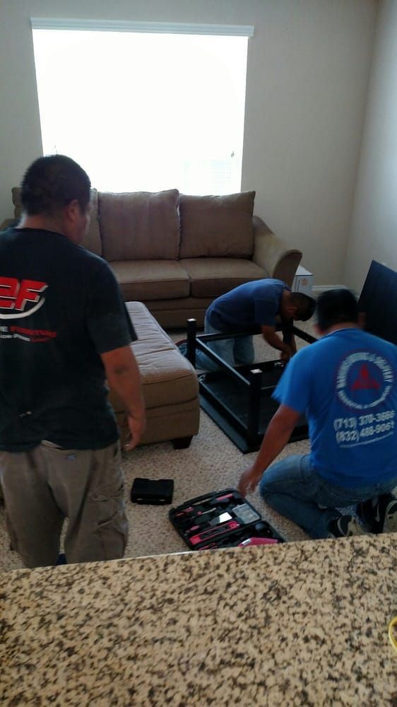 Two people unpack a boxed item in a living room with a sofa and bright window.
