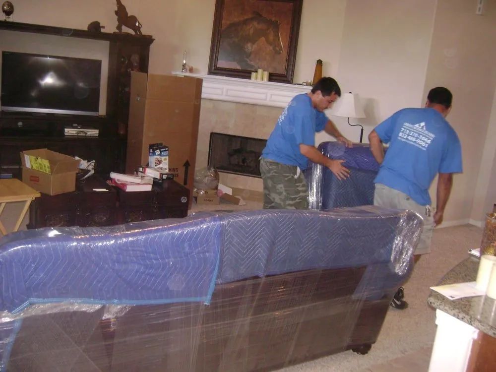 Two men moving a plastic-wrapped couch in a living room with a fireplace and TV.