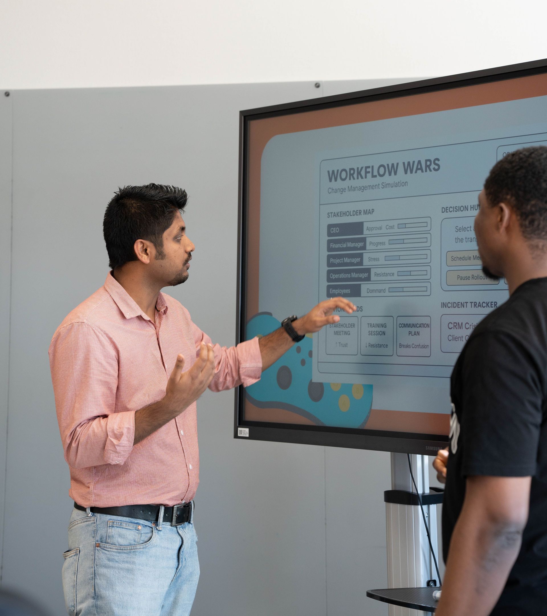 Two men discussing a workflow poster on a wall-mounted screen in an office.