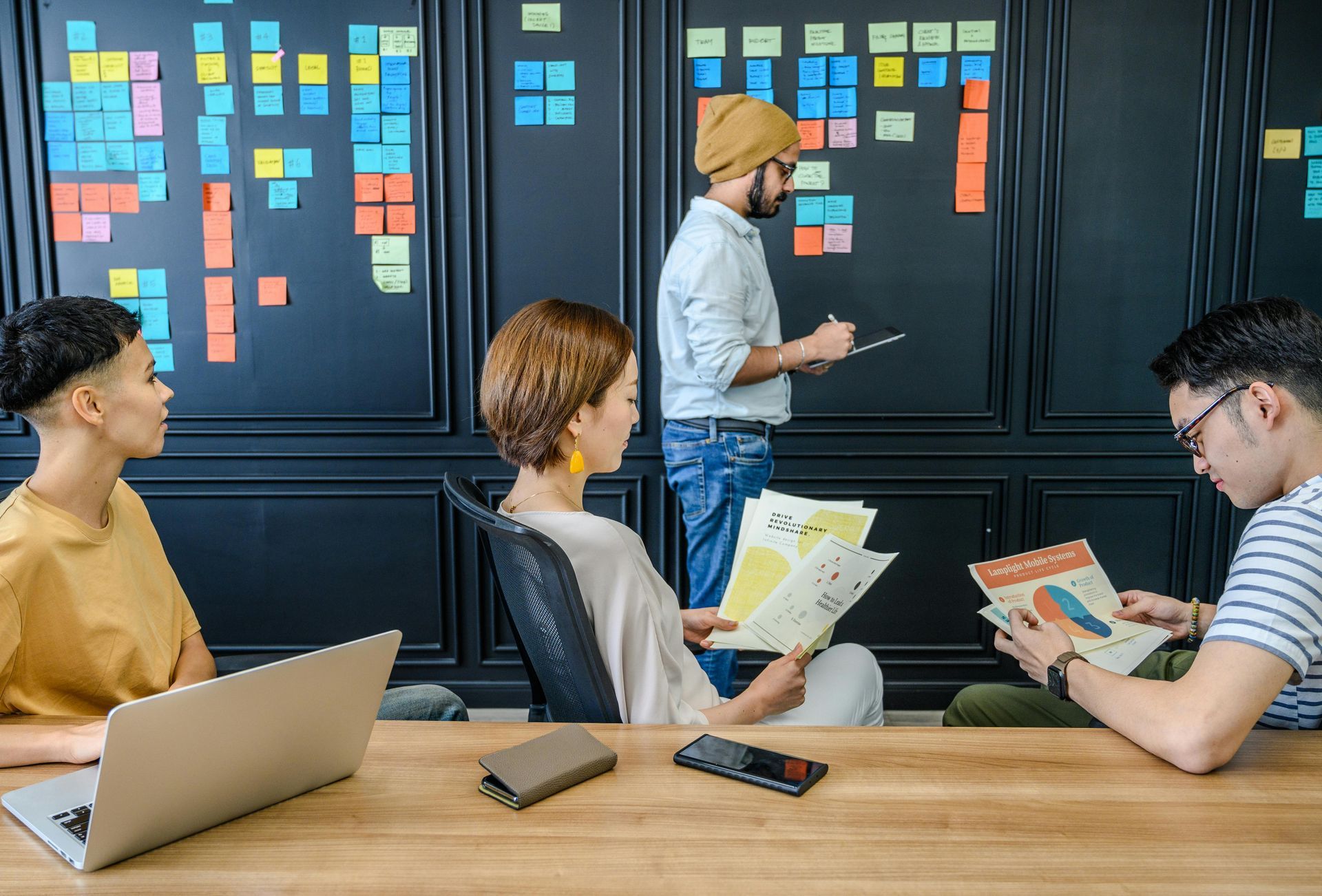 People collaborating in a meeting room with colorful sticky notes on dark cabinets and laptops on the table