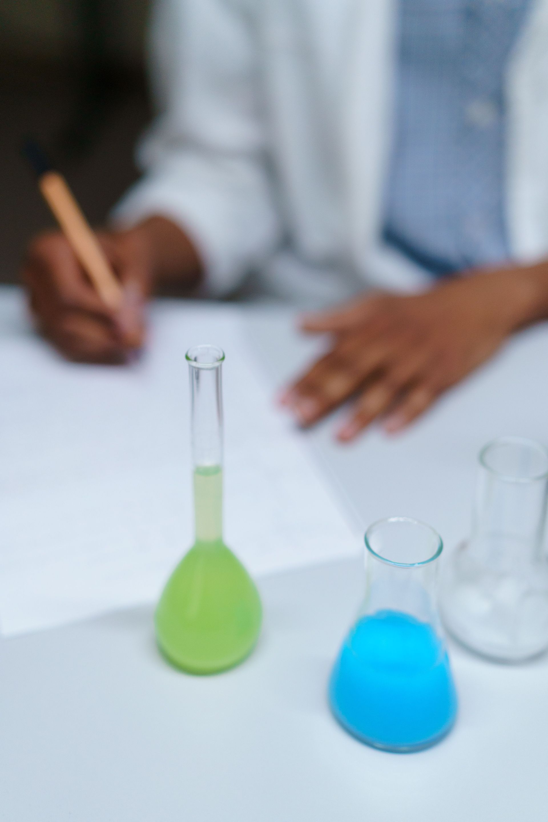 Person in a lab coats holding a pencil near green and blue lab flasks on a table