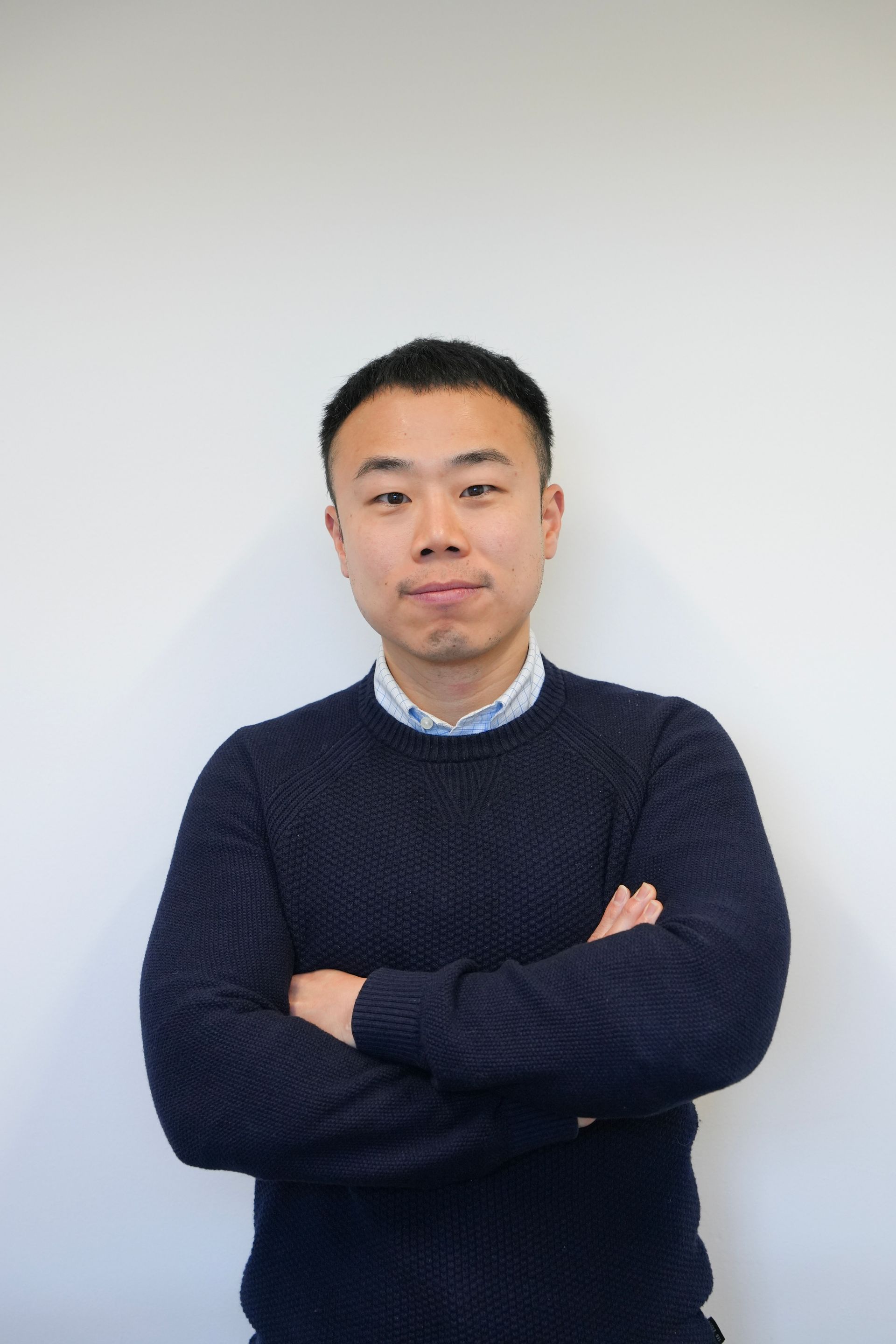 Man in navy sweater with arms crossed, standing against a white wall.