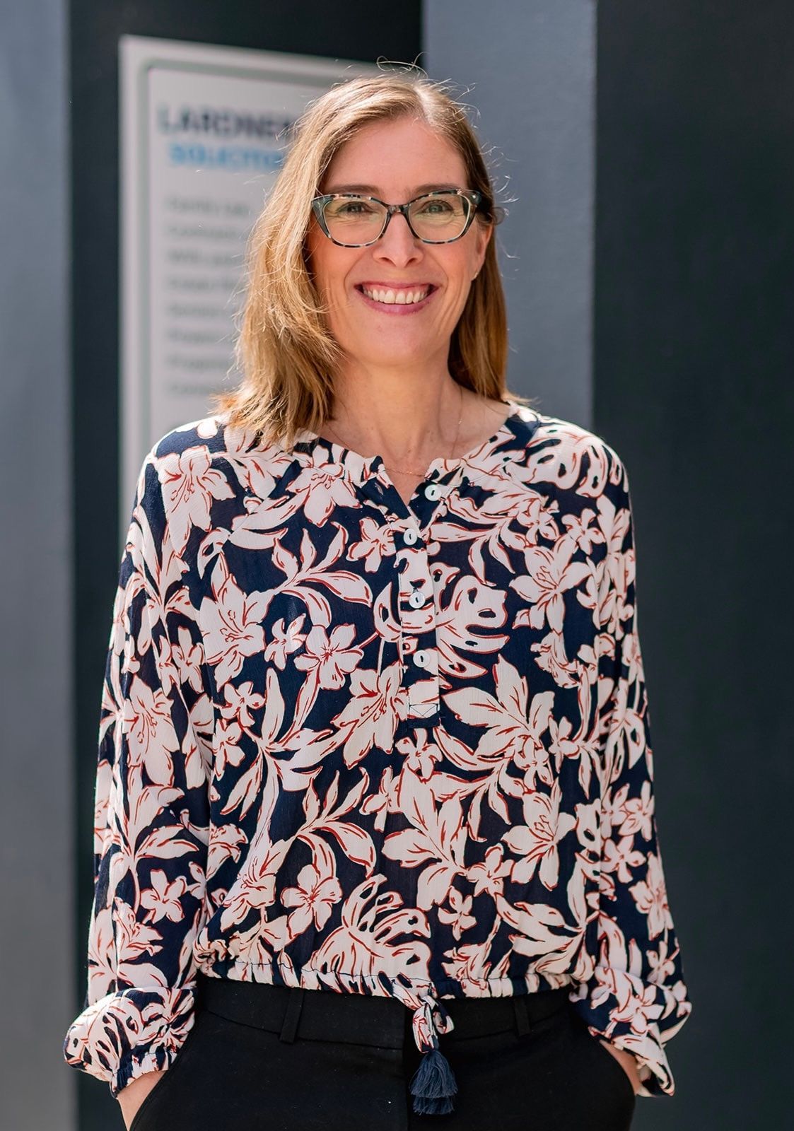 Woman in glasses, patterned blouse, smiling, hands in pockets, standing near a wall with text.